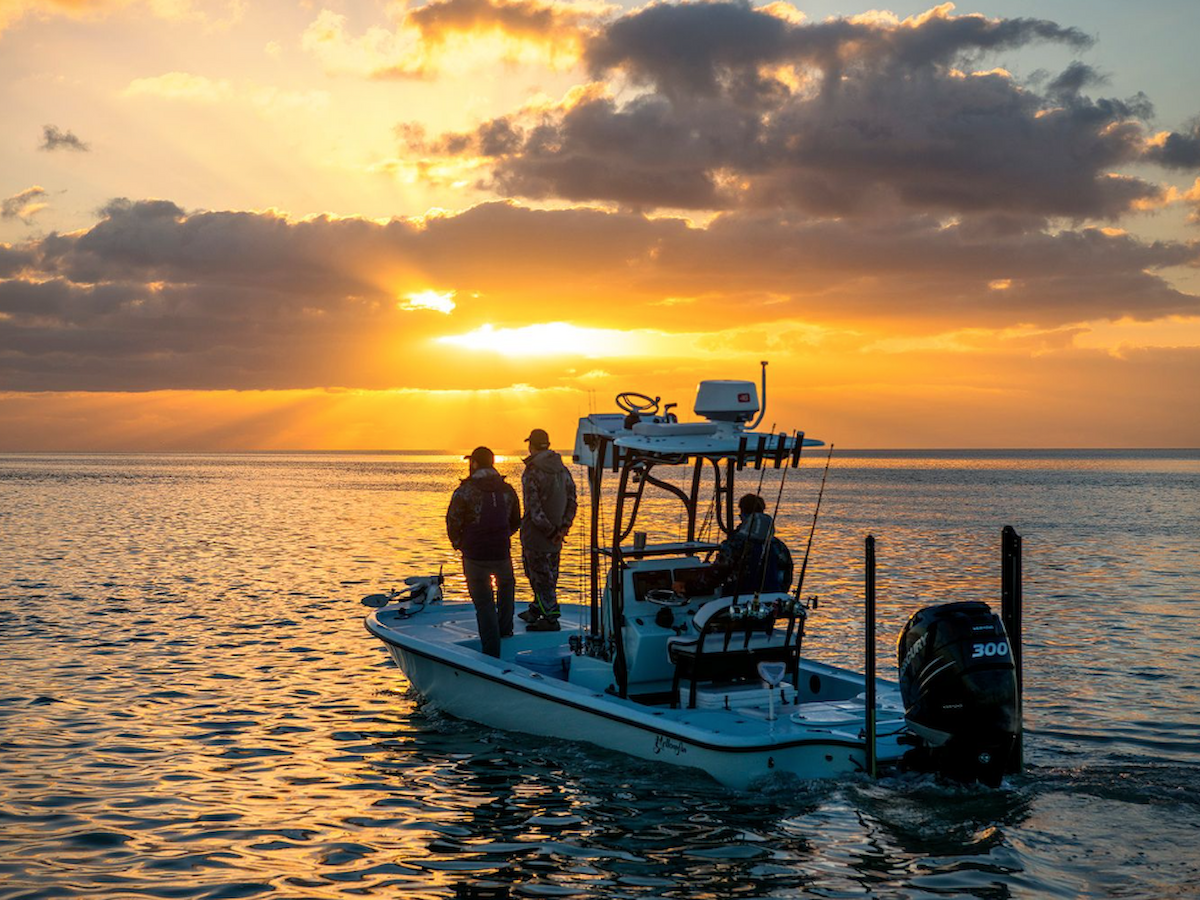 A small fishing boat with three people is on a calm body of water at sunset, with the sun casting a golden glow through the clouds.