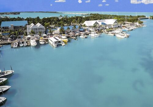 A marina with numerous boats docked, surrounded by clear turquoise waters and coastal buildings under a partly cloudy sky, ending the sentence.