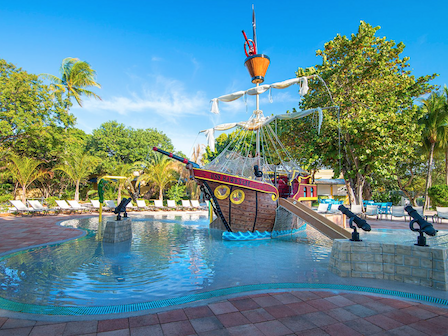 A children's water play area featuring a pirate ship, slide, and canons surrounded by lush greenery and lounge chairs.