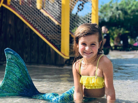 A young girl with a mermaid tail smiles while sitting in shallow water at a water park with play structures in the background.