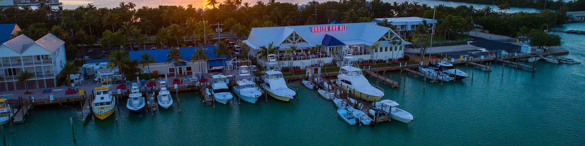 A stunning sunset over a marina with boats docked, surrounded by tropical buildings and palm trees, creating a serene coastal scene.