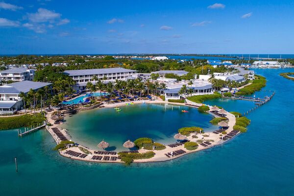 Aerial view of a resort with a lagoon, surrounded by buildings, palm trees, and lounge chairs near clear blue water.