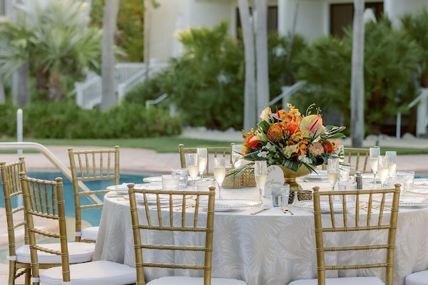 An elegant outdoor dining setup with a round table, gold chairs, floral centerpiece, near a pool, and surrounded by tropical greenery.