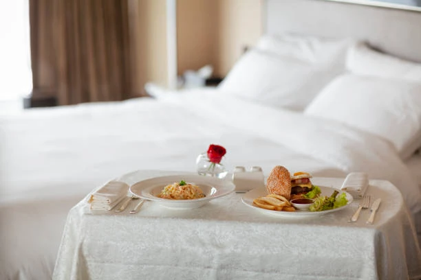 A hotel room with a bed and a table set with food, including a burger, pasta, and a small vase with a red flower.