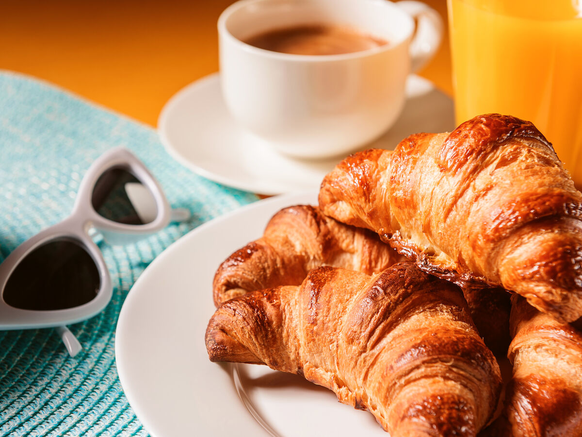 Plate of croissants, cup of coffee, glass of orange juice, sunglasses, and a blue placemat on a table.